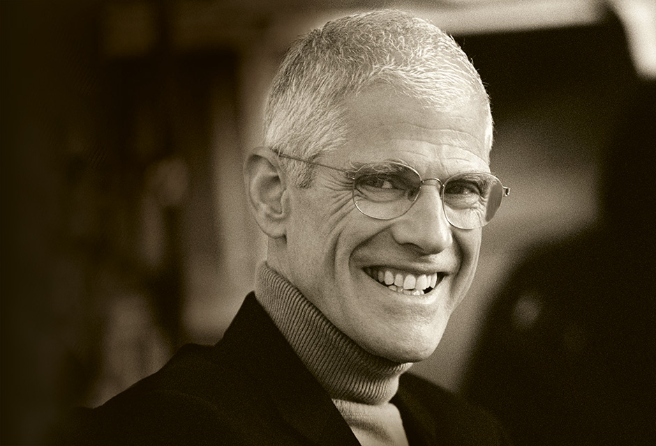A black and white headshot of Sir Peter Jonas, who is wearing a roll-neck jumper, a blazer and glasses, and smiling at the camera.